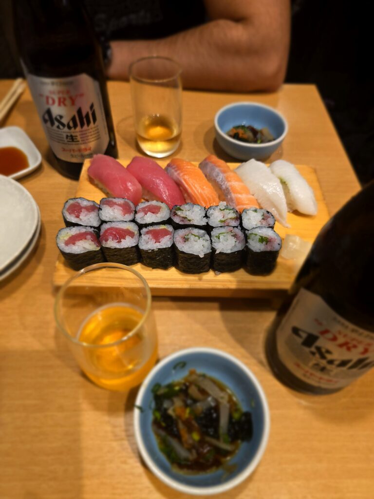 bandeja de madera con una variedad de sushi (nigiris de atún, salmón y pescado blanco, y makis) acompañada de cervezas Asahi Super Dry y cuencos pequeños con comida.