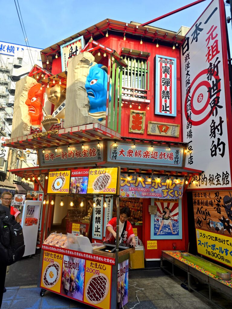 colorida y llamativa fachada del complejo de entretenimiento y galería de tiro japonesa (shateki) Hihoukan (con una de sus sucursales principales conocida como la 秘宝館 本店 o Hihokan Main Store) ubicada en el animado barrio de Shinsekai, justo al lado de la famosa torre Tsūtenkaku en Osaka, Japón