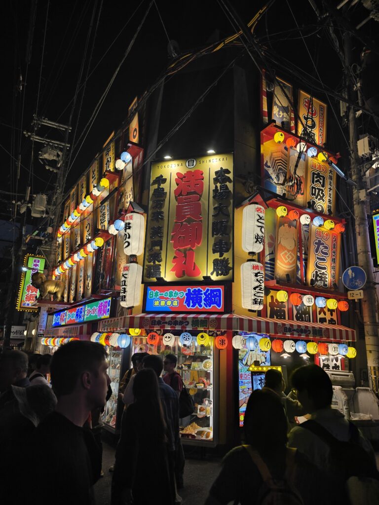 esquina nocturna en el distrito de Shinsekai, en Osaka (Japón), caracterizada por sus llamativos carteles luminosos, farolillos de papel y un ambiente de estilo retro.