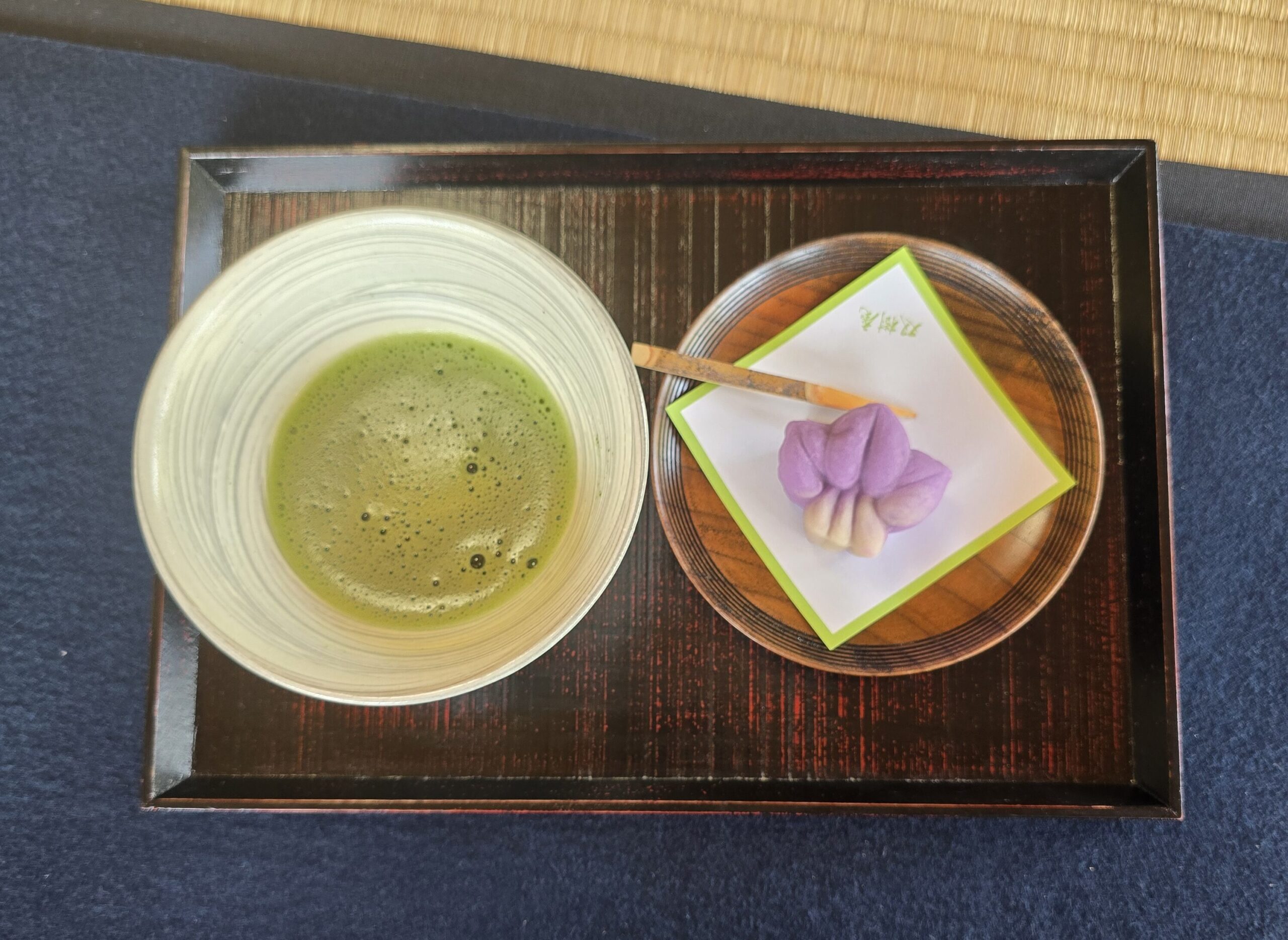 bandeja de madera con un cuenco de té matcha espumoso a la izquierda y un dulce japonés tradicional (wagashi) de color morado y blanco colocado sobre un plato pequeño con un palillo a la derecha.