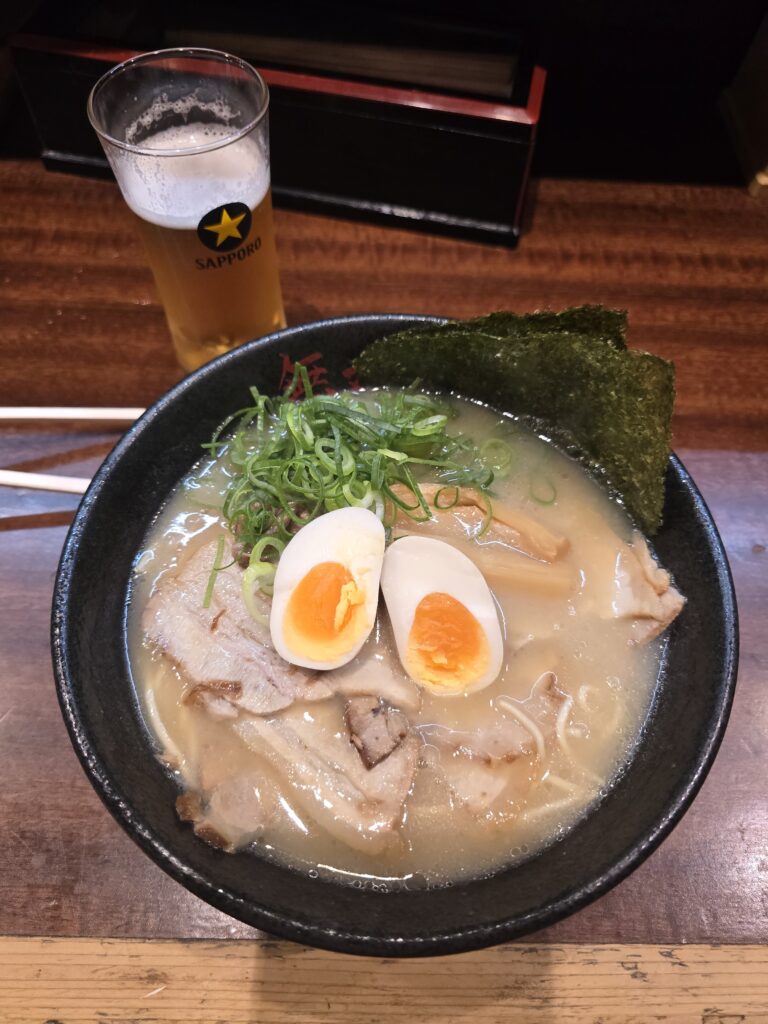 ramen con fideos, caldo claro, rebanadas de cerdo chashu, un huevo ajitsuke partido por la mitad, cebollino picado y alga nori, acompañado por un vaso de cerveza.