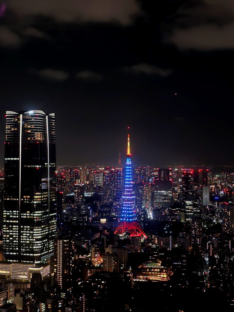 Torre de Tokio (Tokyo Tower) iluminada de noche en el barrio de Minato, flanqueada a la izquierda por un gran rascacielos moderno.
