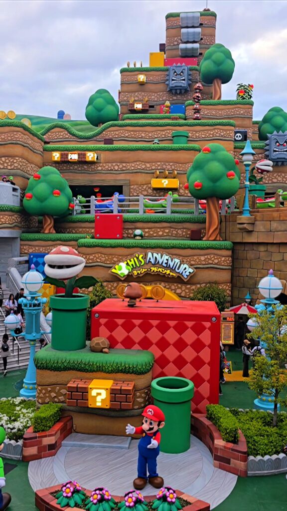 Decoración colorida de Super Nintendo World con tuberías verdes, plantas piraña, bloques de interrogación y el letrero de la atracción Yoshi's Adventure.