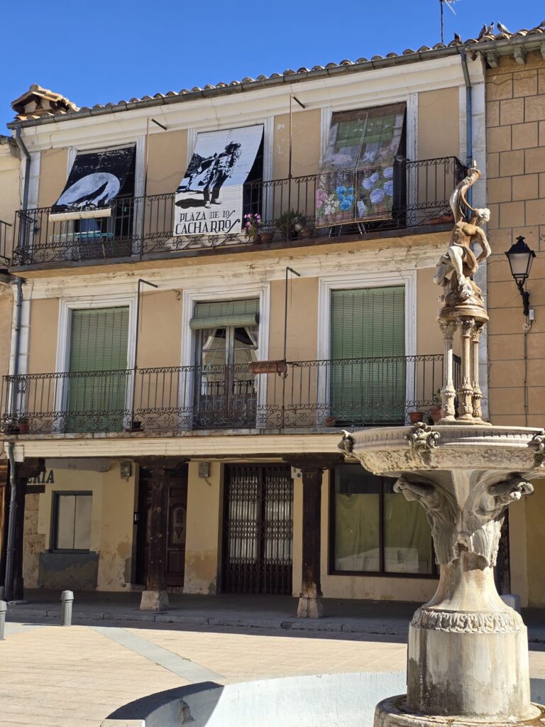 Detalle de una plaza histórica en El Burgo de Osma, mostrando balcones con persianas verdes, soportales de madera y una fuente con una escultura en primer plano.