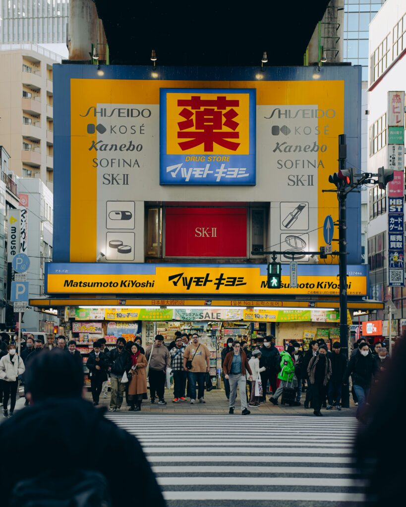 concurrida calle de Tokio, Japón, con una gran fachada amarilla y azul de una tienda de la cadena de farmacias y cosméticos Matsumoto Kiyoshi, destacando el característico kanji "薬" (medicina) en la parte superior
