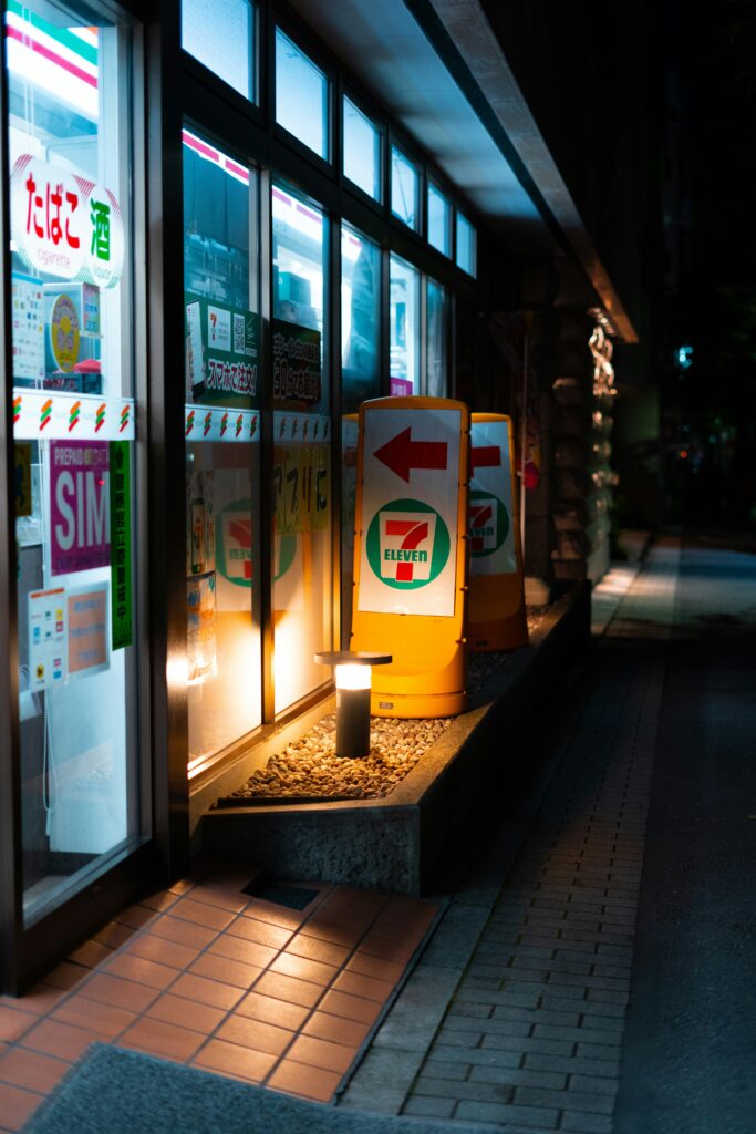 Tienda de conveniencia japonesa (konbini) 7-Eleven iluminada de noche, mostrando un cartel tradicional de tabaco y alcohol junto a un letrero de tarjeta SIM para turistas.